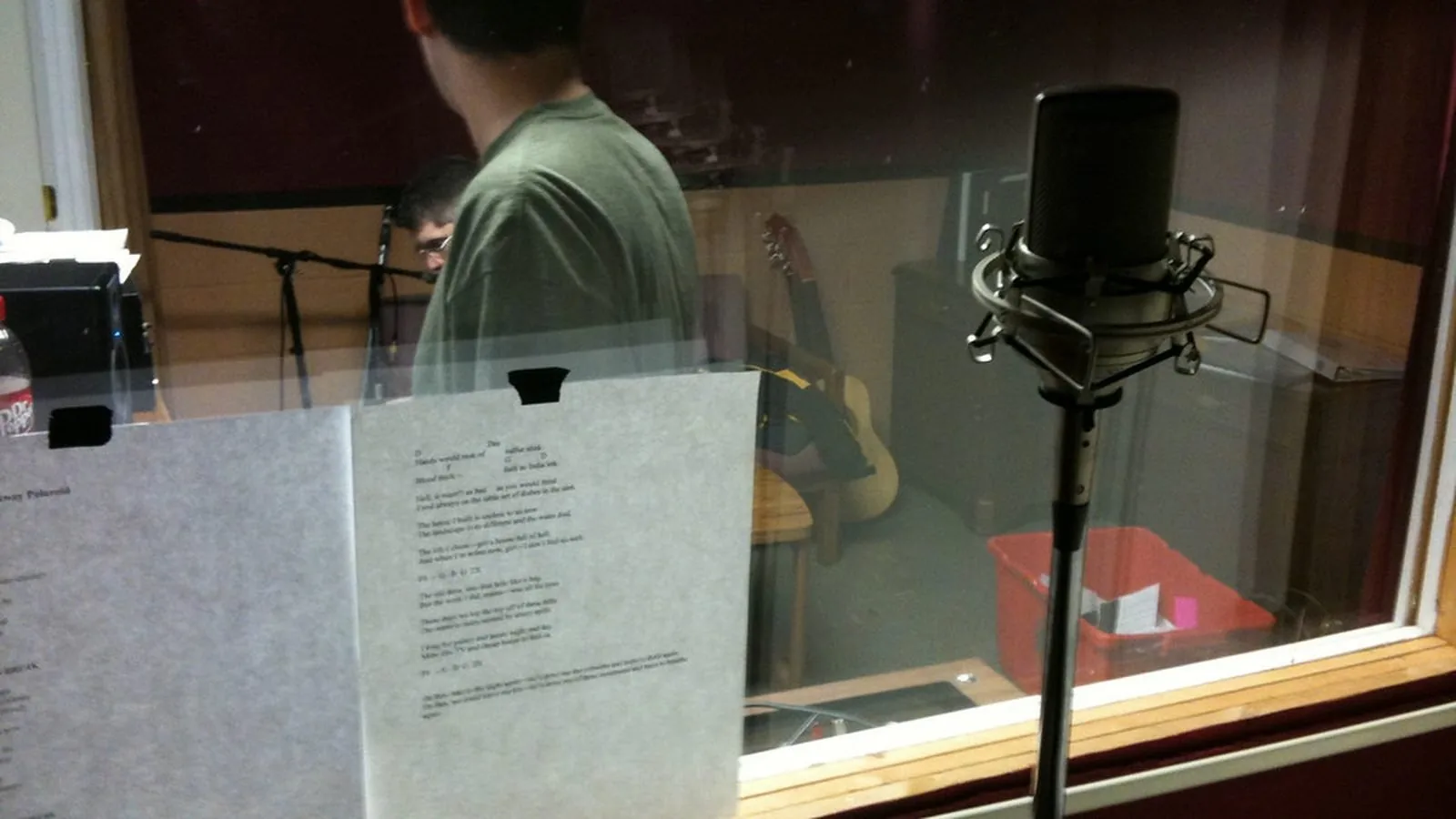 A person seated inside a sound booth, seen through the observation window